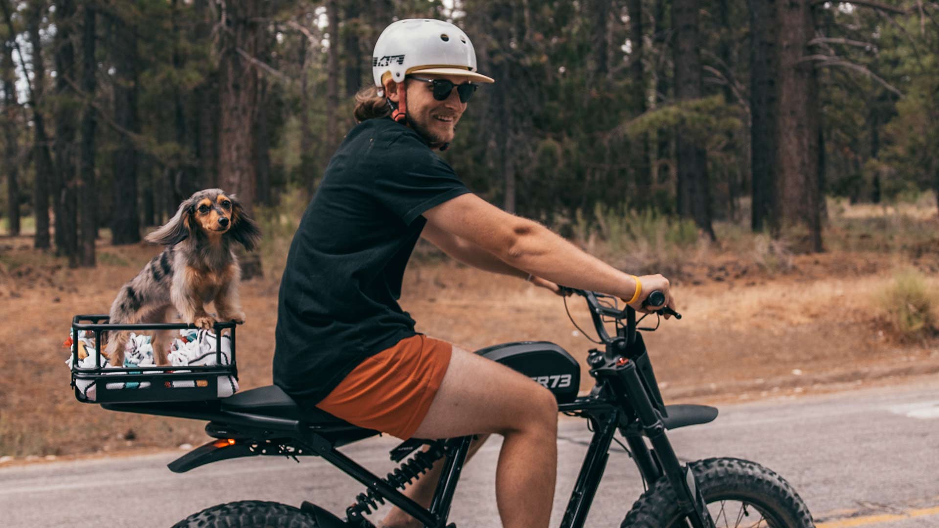 Young man riding a Super73 ebike on the road with a rear rack attached and his dog in a basket